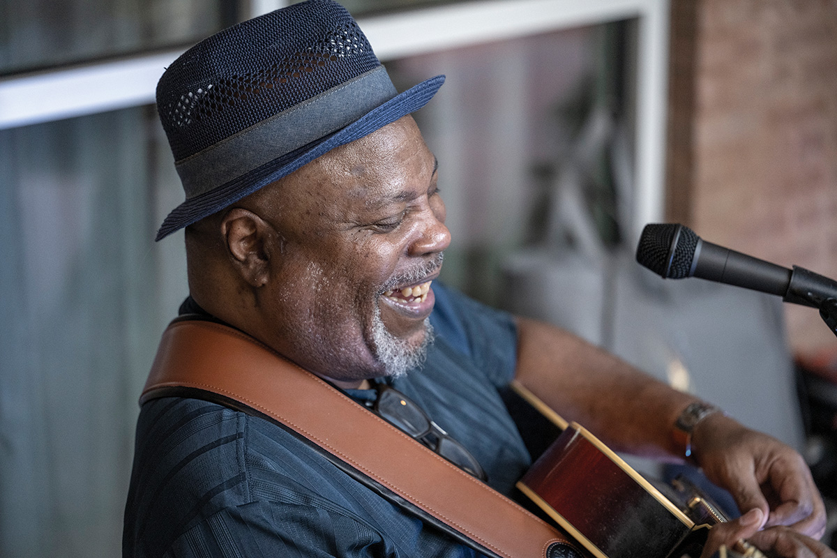 A man with a goatee, shaved head, hat sits and plays guitar, smiling