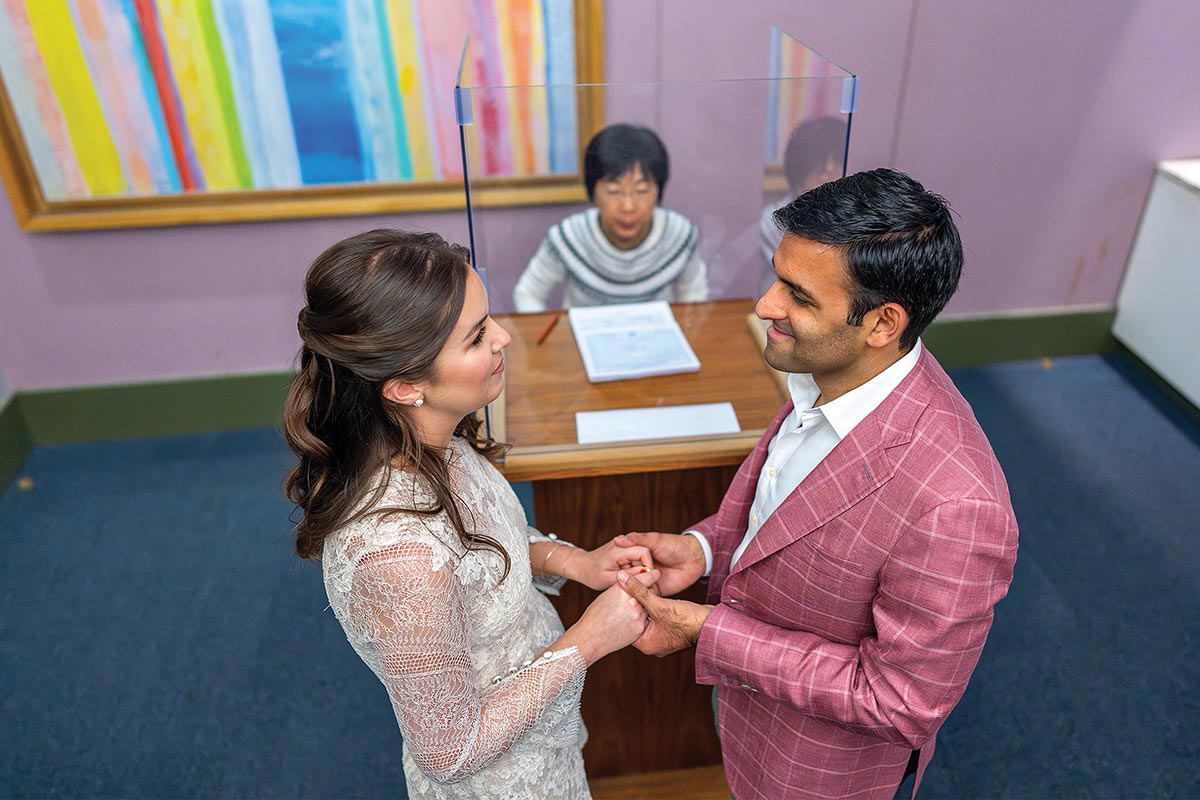 A bride and groom stand holding hands in front of the city clerk to take their vows. The room is shabby with pink walls, dark green trim, and worn blue carpet. The clerk is behind a plexiglass shield.