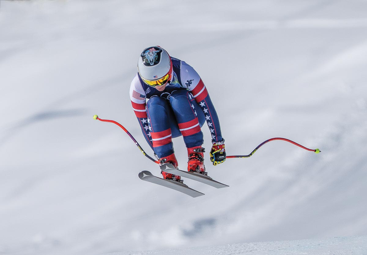 A downhill Alpine skier in mid-air, all-snow background, knees-to-chest poles-down position, red, white, and blue ski suit