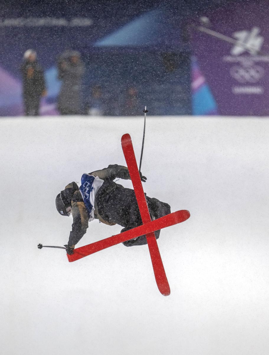 A big air ski competitor in the Winter Olympics is in mid air, looking down as he twists, his red skis crossed in an X shape