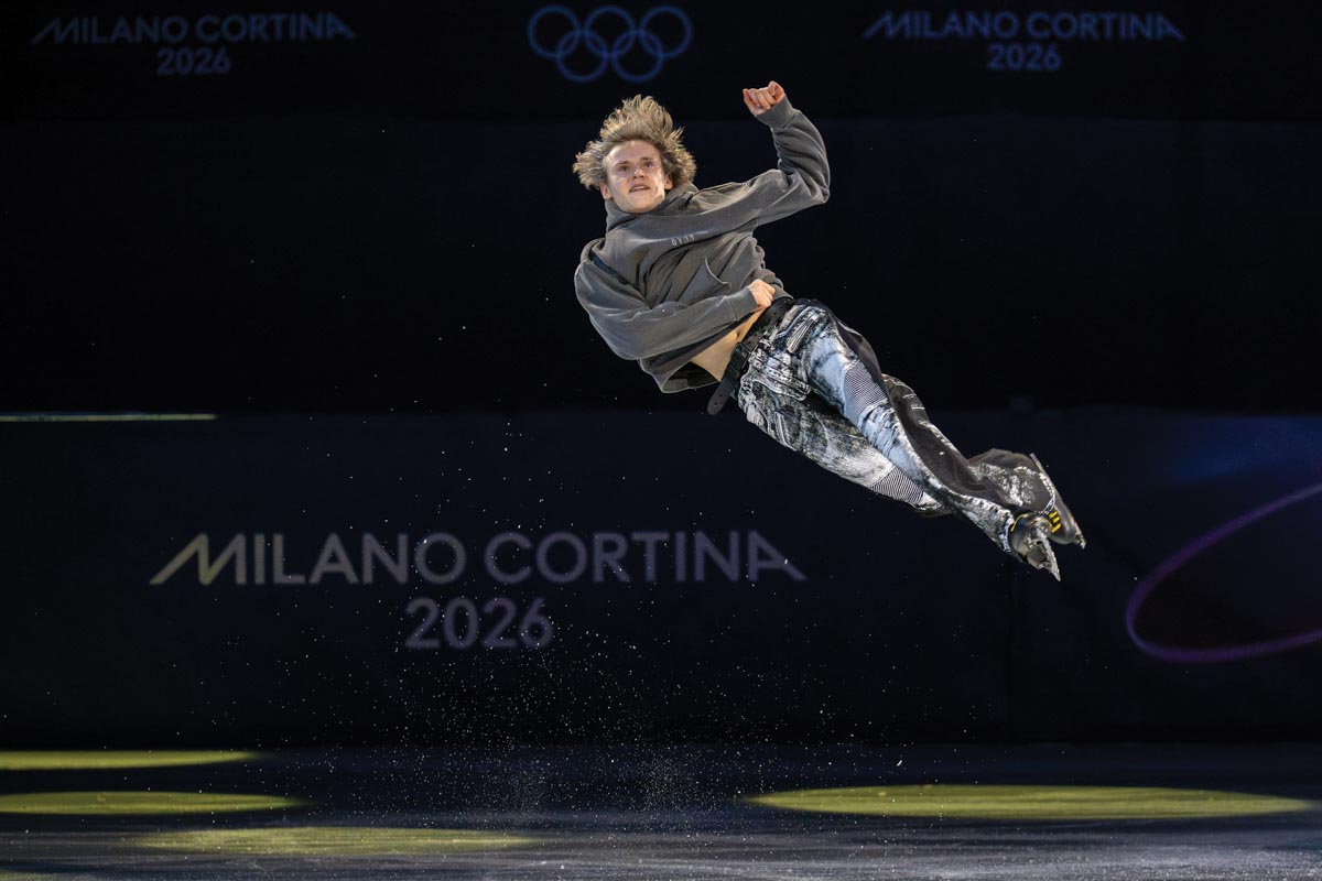A white male figure skater in a loose gray sweatshirt and black bleach-patterned jeans is frozen in a leap in which he is at about a 50-degree angle, right arm clenched to his torso and left arm up, ankles crossed; the background is mostly black