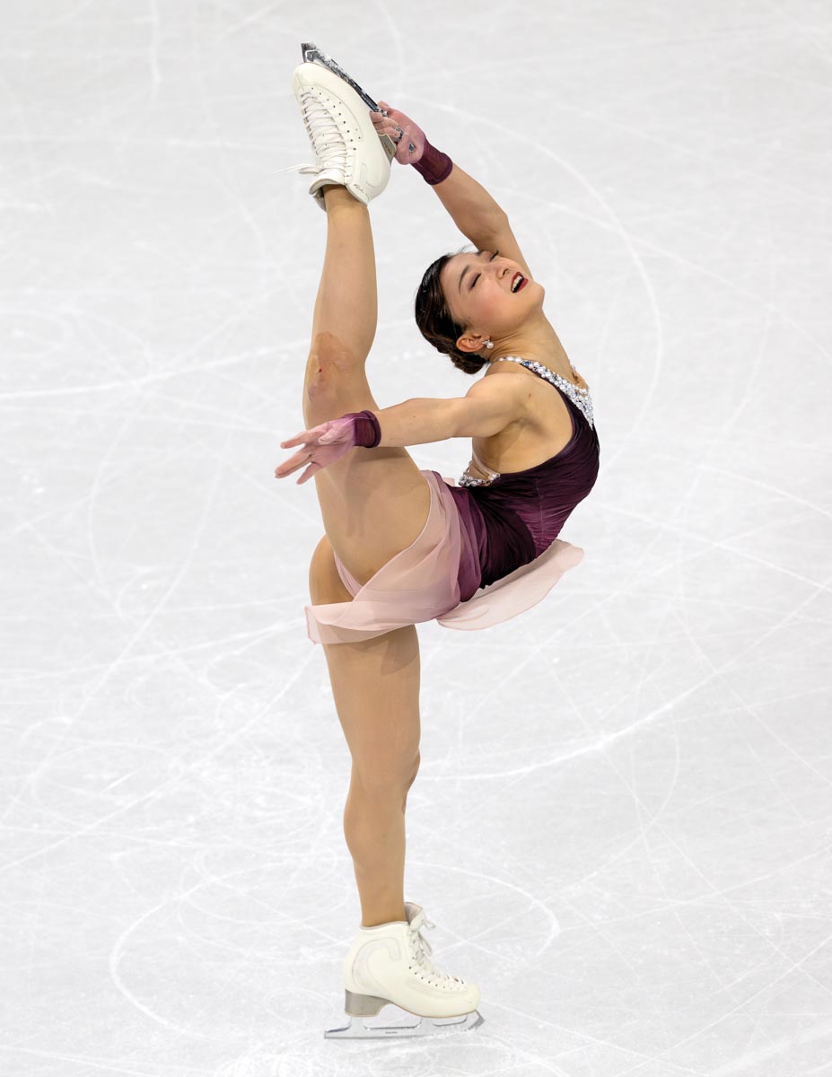 a woman figure skater, frozen in action, her left hand extended above her head holding the blade of her right skate as her legs do a vertical split; her costume is maroon and pink