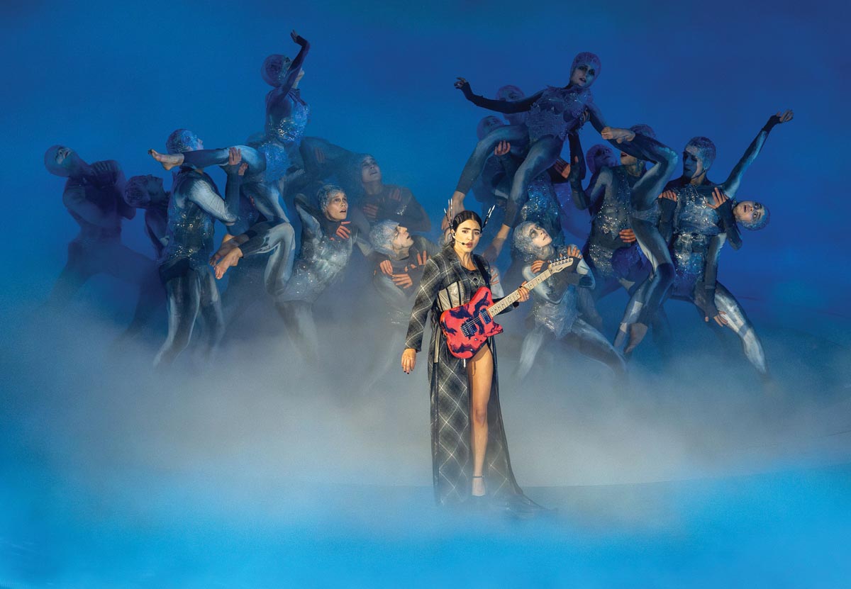 An Asian woman in a floor length diamond-patterned robe, with a headset, holding a red guitar, sings in front of a group of dancers against a blue background with fog surrounding them