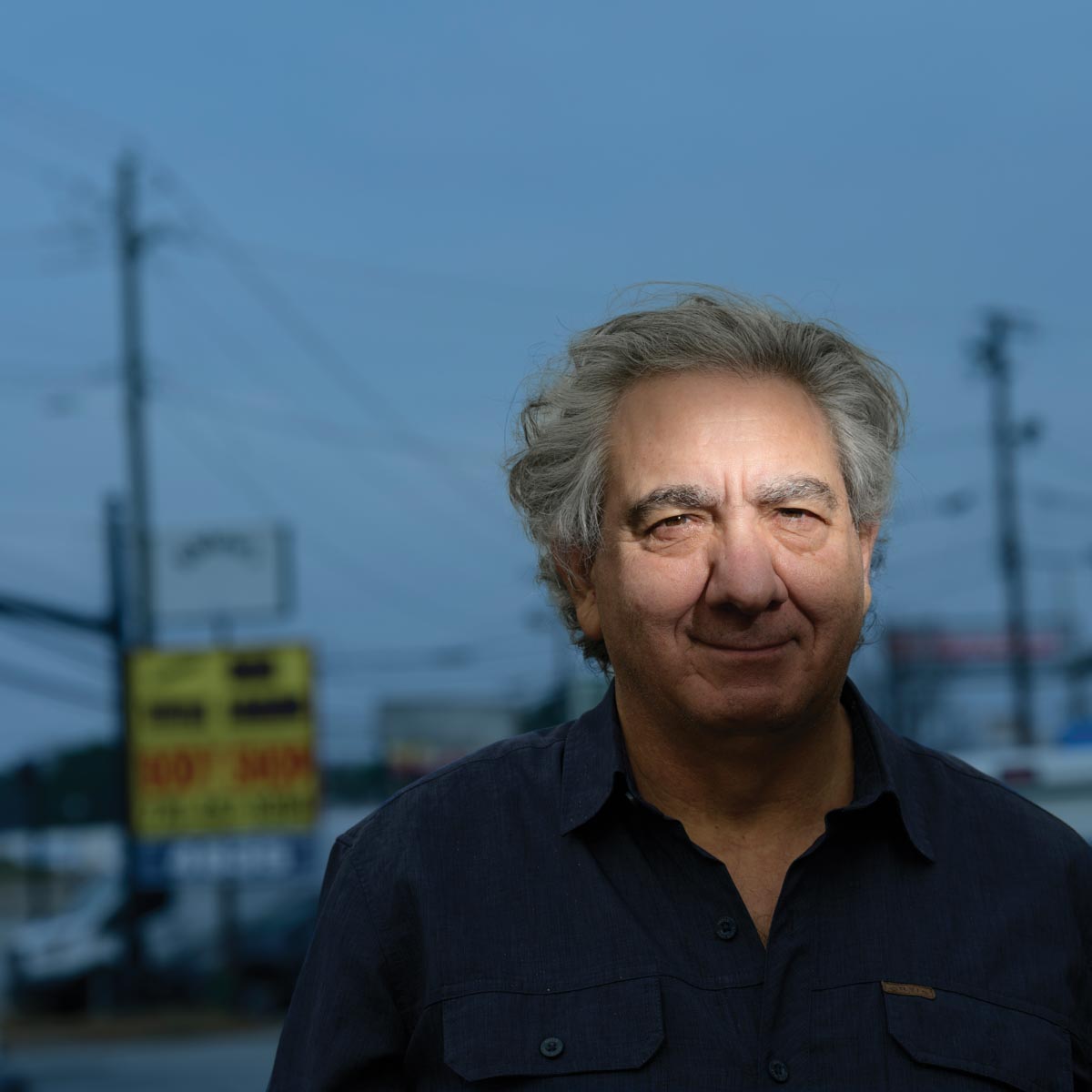 An older white man outside at dusk, slightly dark blue sky with power lines and signs out of focus behind him. He’s wearing a black shirt and only the top of his face is lit with flash. 