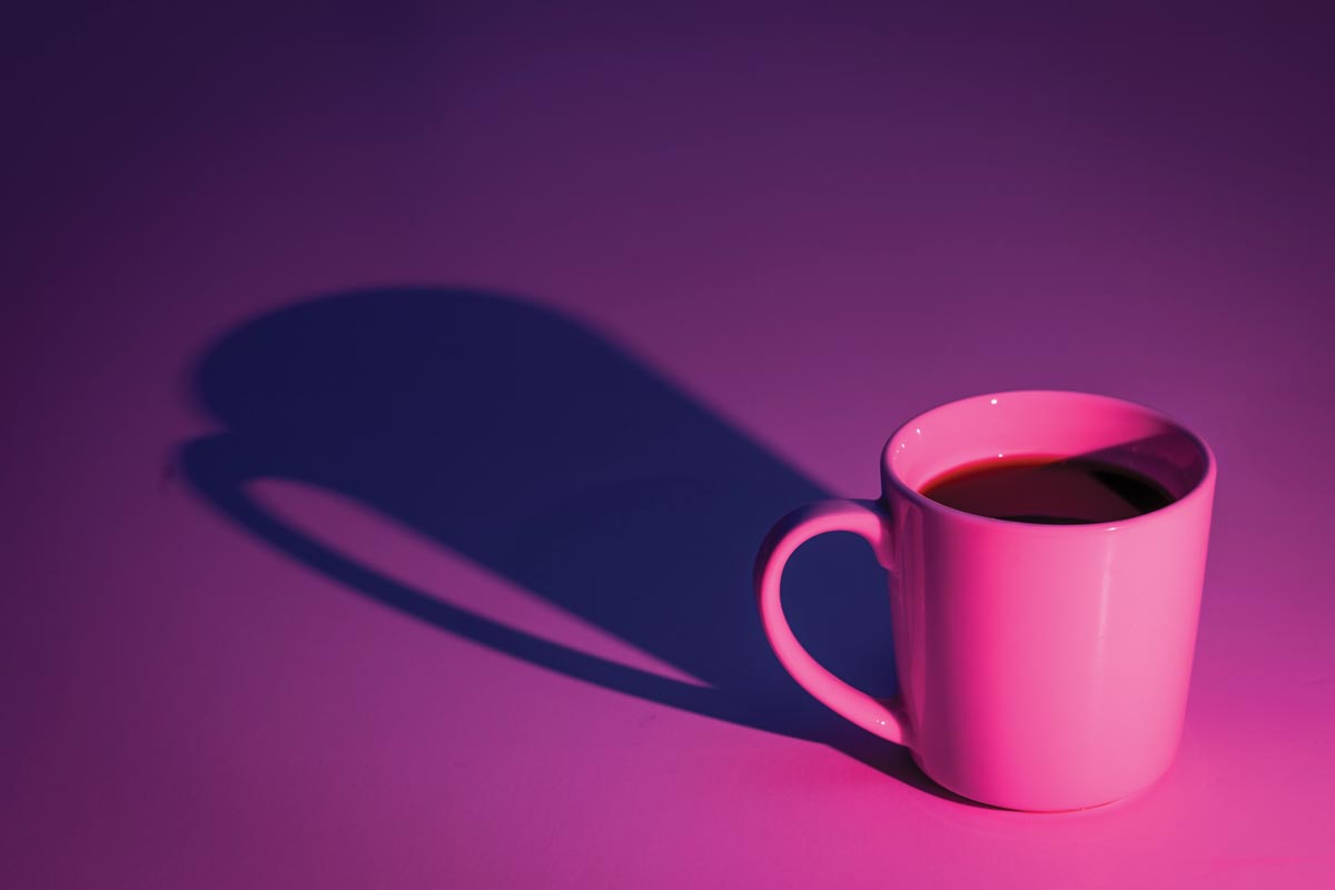 A white coffee mug sits on a white background, but the scene is lit with a blue gelled light, and a tangerine gelled light. The mix makes the background a deep purple, and the coffee mug looks pink. The directional light casts a dramatic mug shadow.
