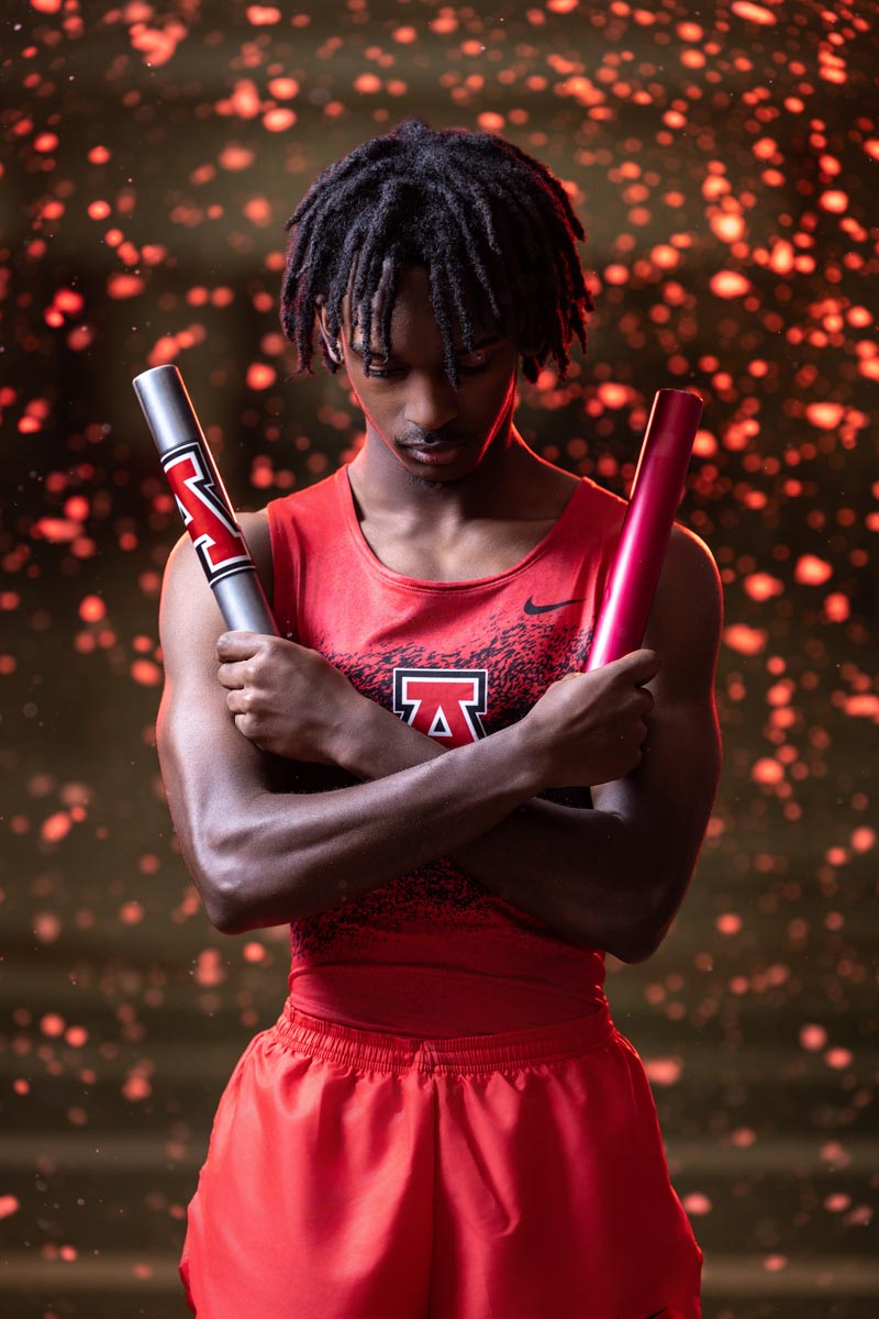 a high school track athlete with arms crossed and head down with two batons in his hands and fake snow that is lit up read in the background
