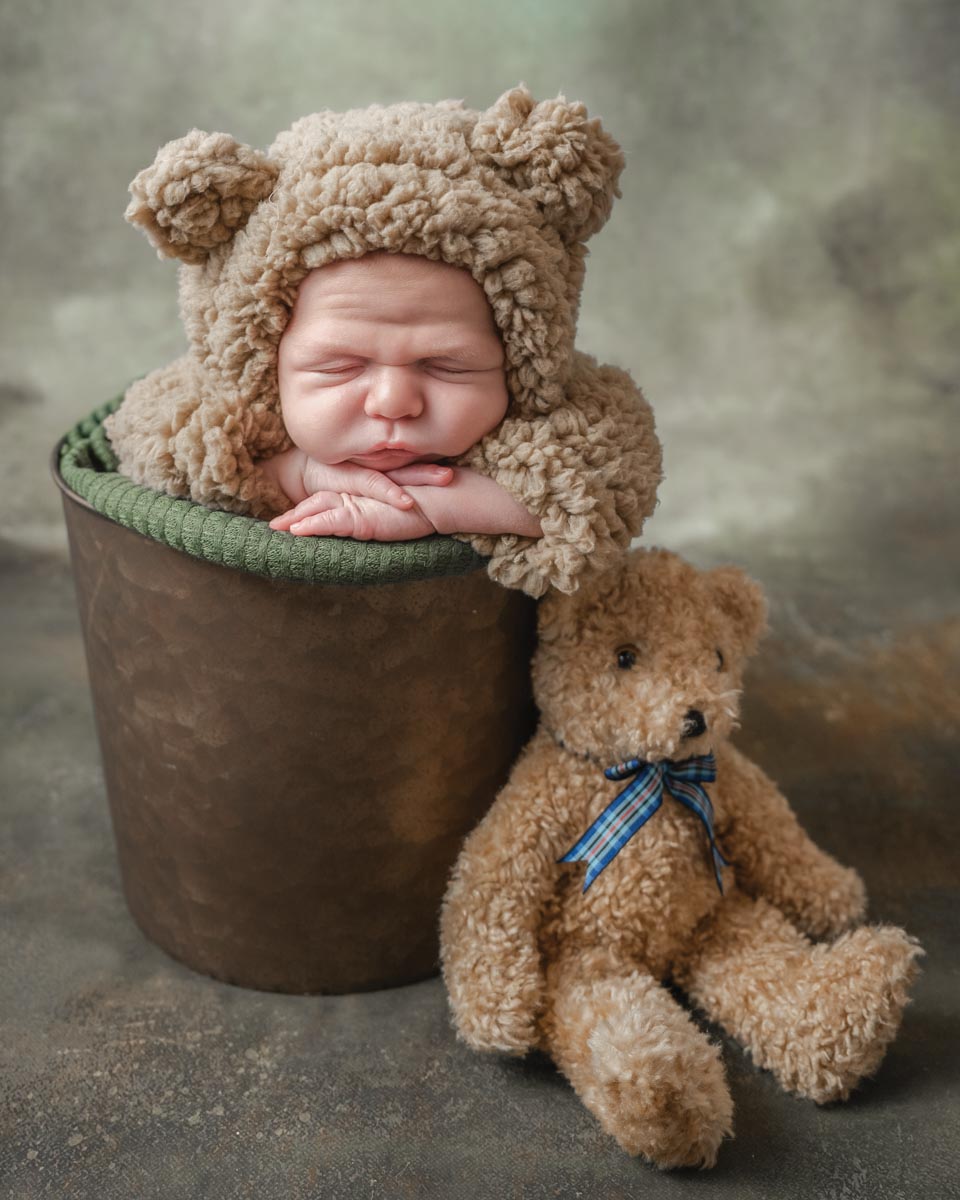 a portrait of a newborn wearing a bear onesie with a hood inside a bucket