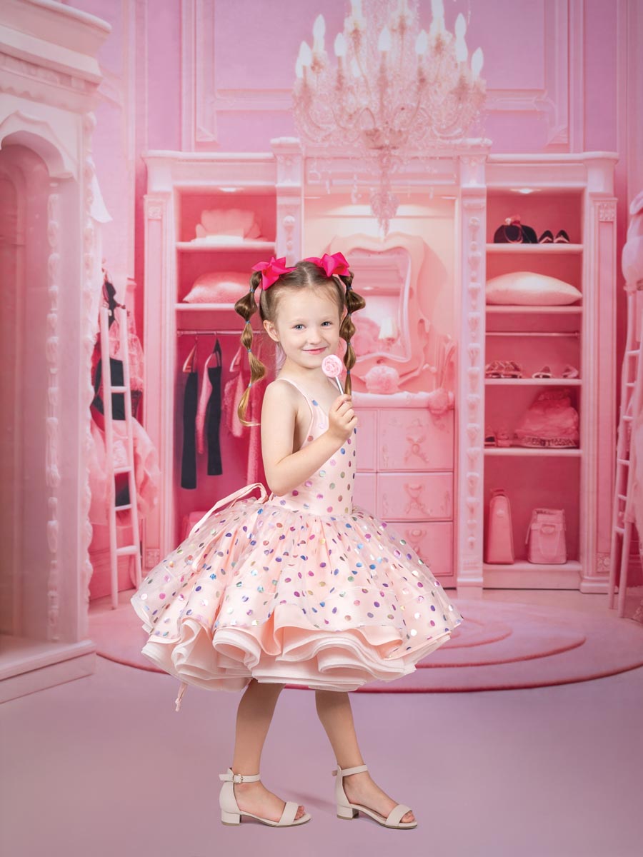 portrait of a young girl holding up a lollipop in a pink dress with pink bows on her braided hair with a pink dressing room backdrop
