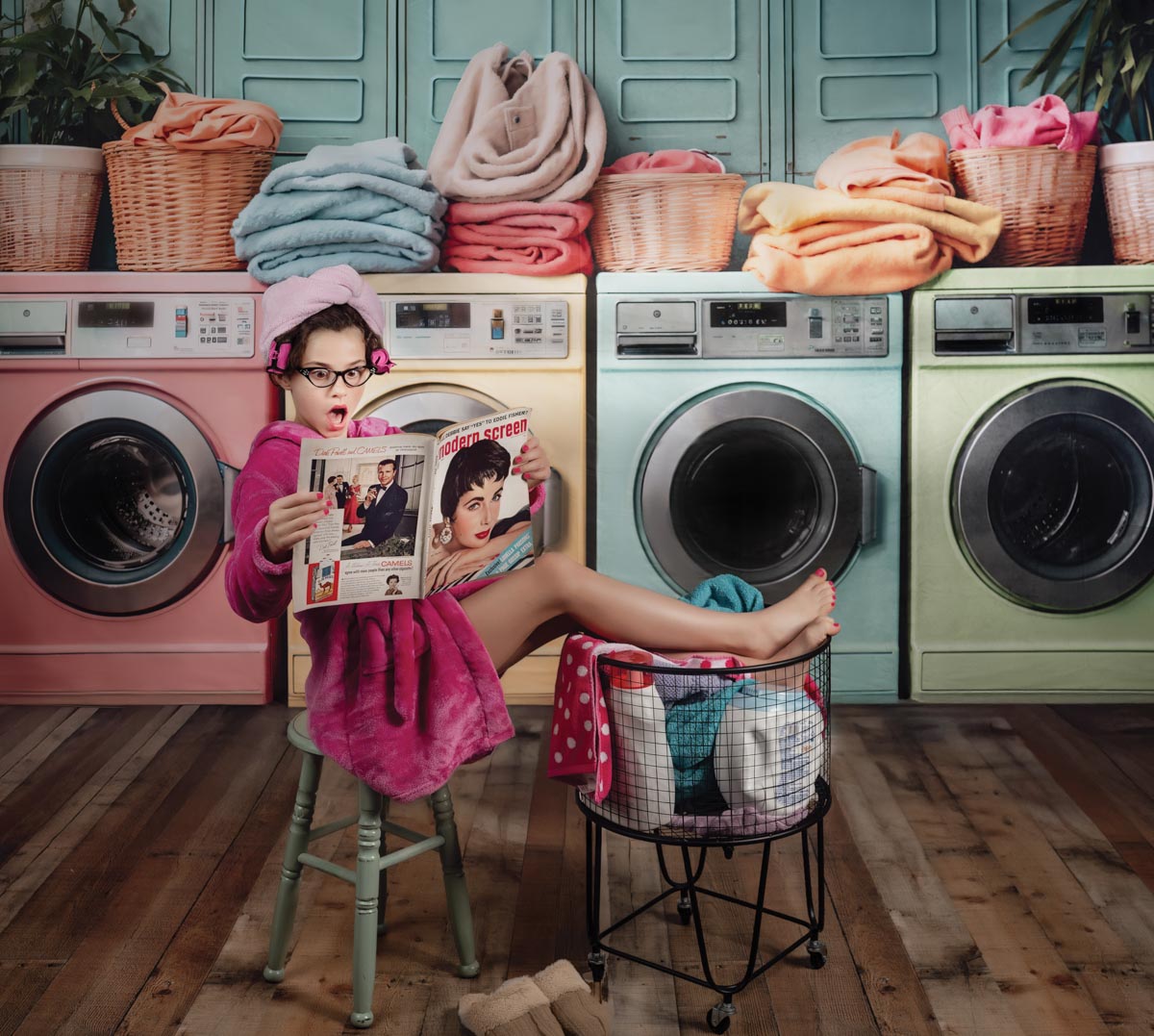 portrait of a young girl sitting in a chair with her feet up reading a magazine with a towel on her head and with a row of washers and piled up laundry behind her