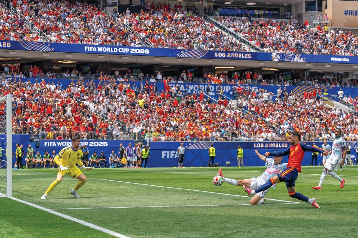 On a 2026 World Cup soccer pitch, a goalkeeper in yellow kit is ready to defend a goal attempt made by a Spain player in red and blue kit. A Belgian player in blue, pink, and white kit is sliding, one leg extended, to block the ball. 