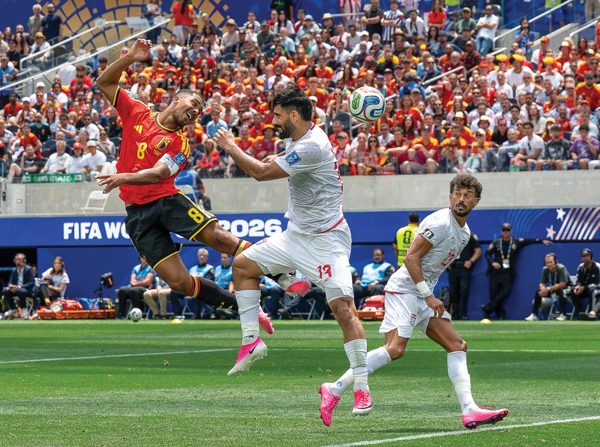 Three soccer players are simultaneously in the air on a World Cup soccer pitch. The Belgium player in a red and blue kit has headed the ball. An Iran player is in the air next to him trying to block, and another Iran player in a white kit is running.