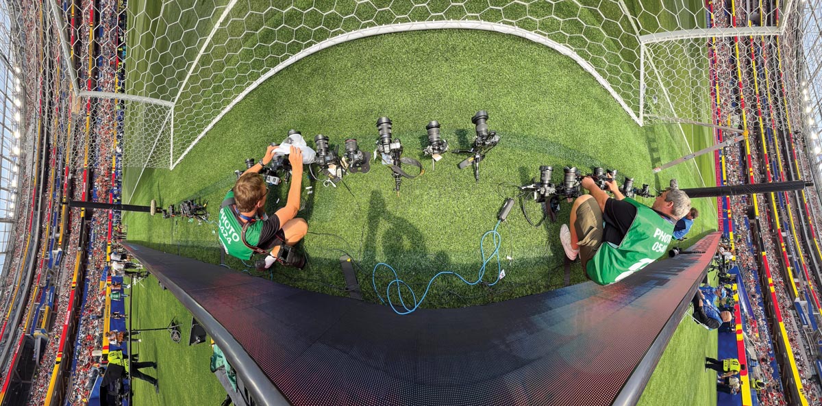 Two photographers in green vests indicating they have credentials are behind a soccer goal setting up remote cameras. Several cameras are already set up between them. 
