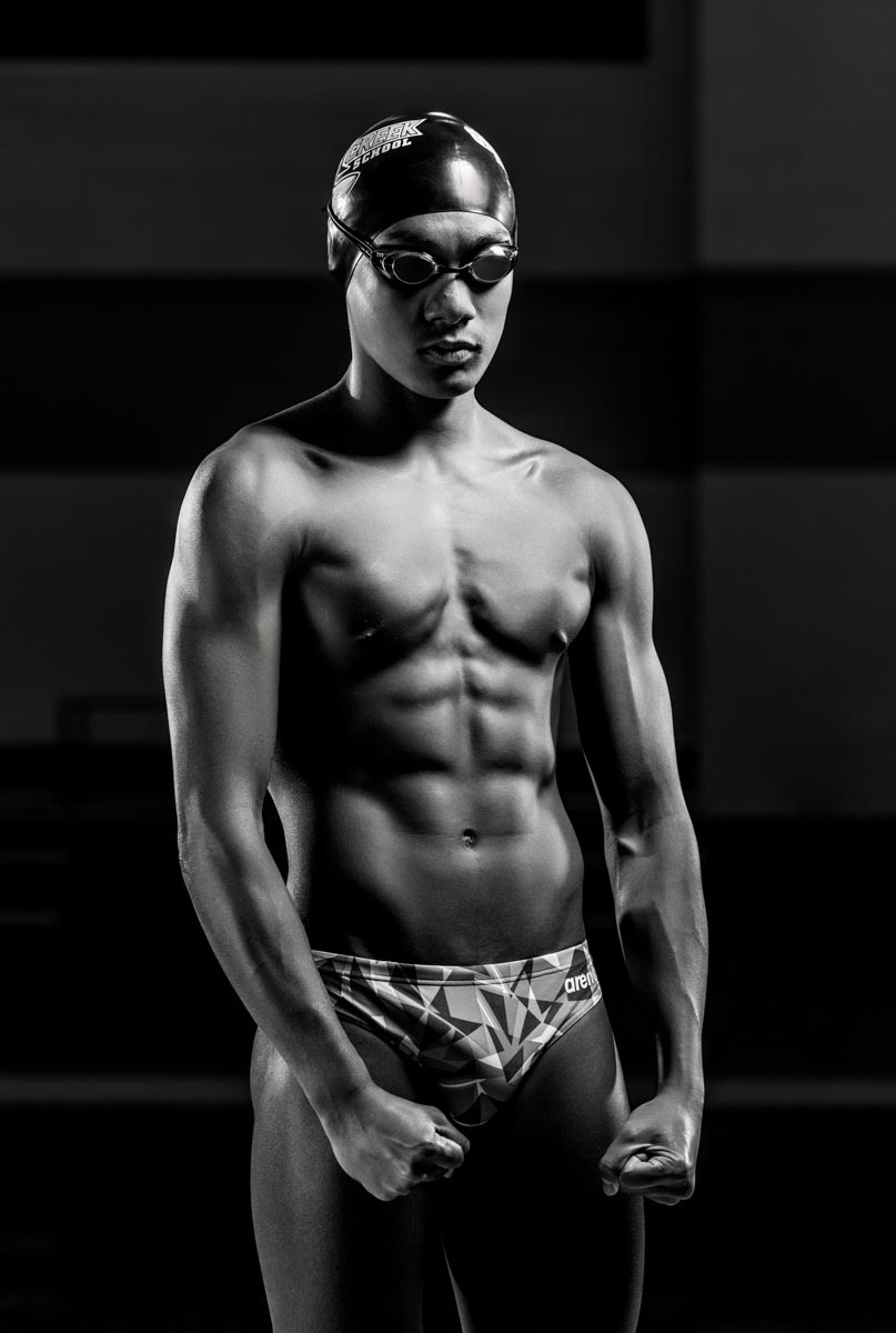 a black-and-white portrait of a male high school swim athlete flexing his muscles and wearing goggles 
