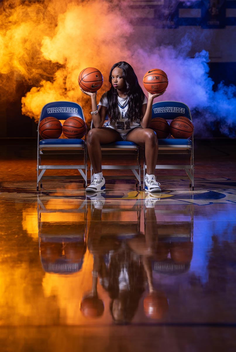 a high school female basketball player sitting in a chair in a gym holding up two basketballs with smoke in the background