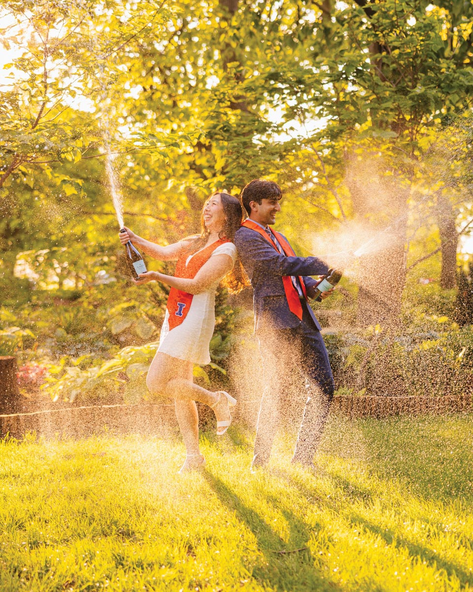 two college graduates back-to-back on a lawn wearing their school colors and spraying champagne into the air