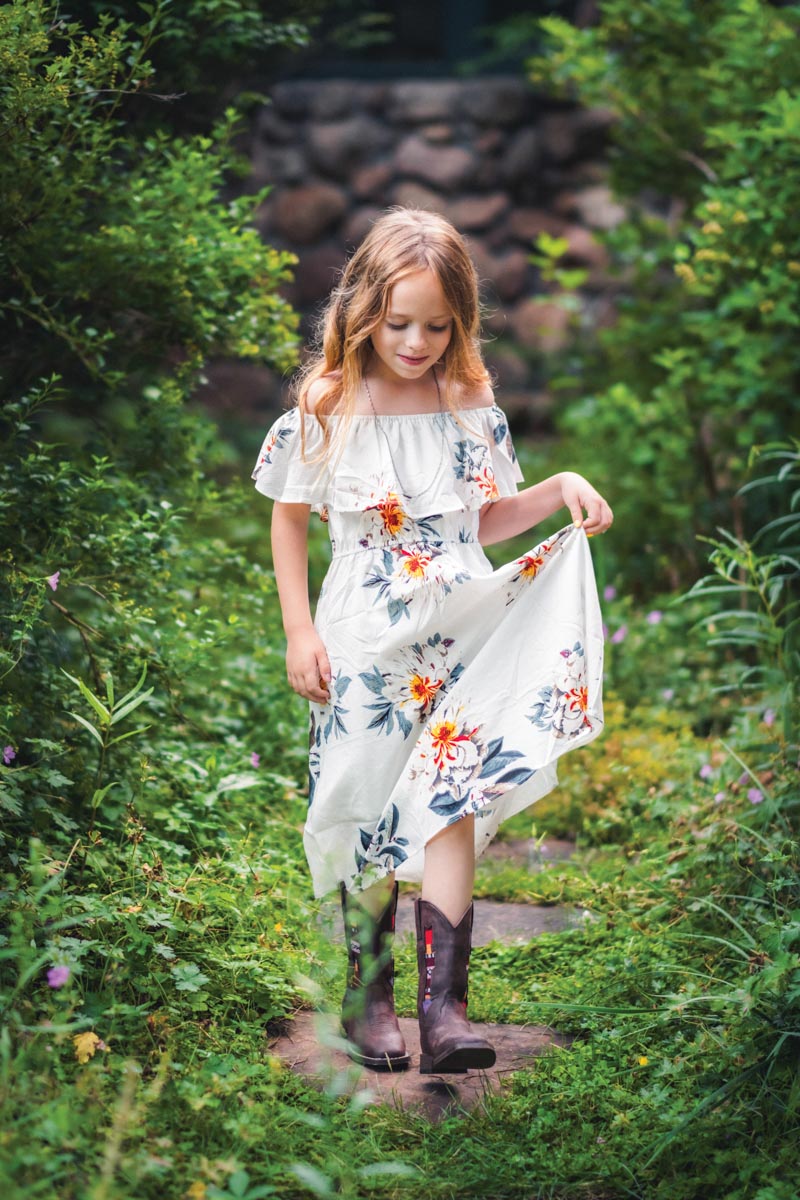 young girl in a white floral dress and cowboy boots walking through a wooded path