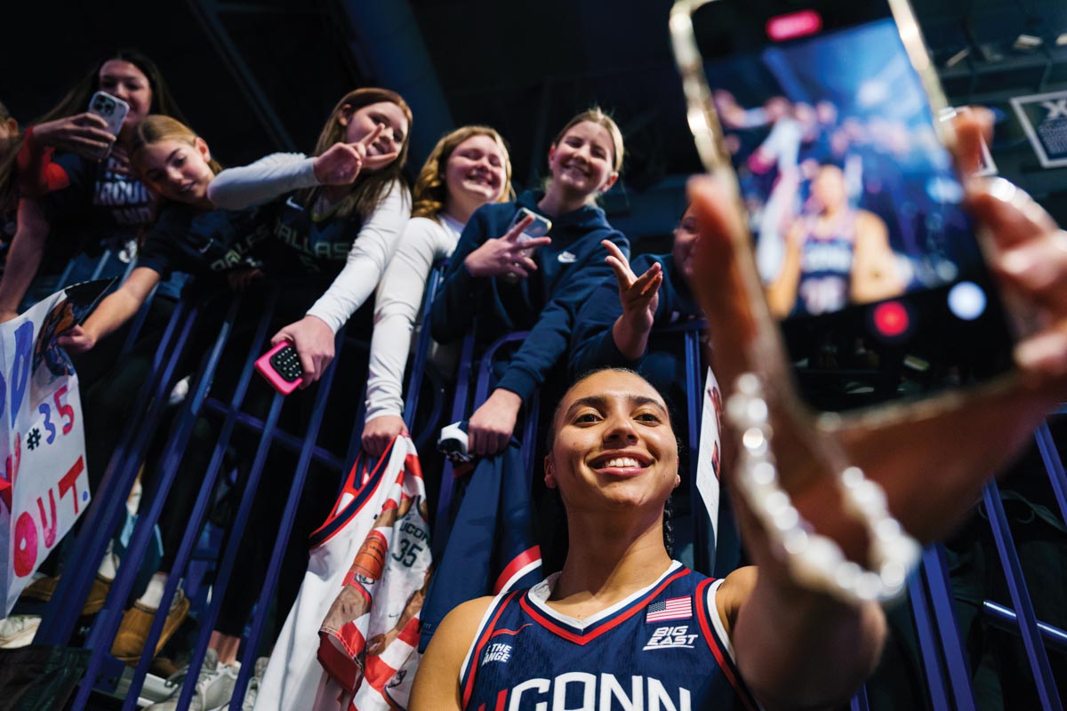 female basketball player holding up a photo on her phone with fans above and behind her