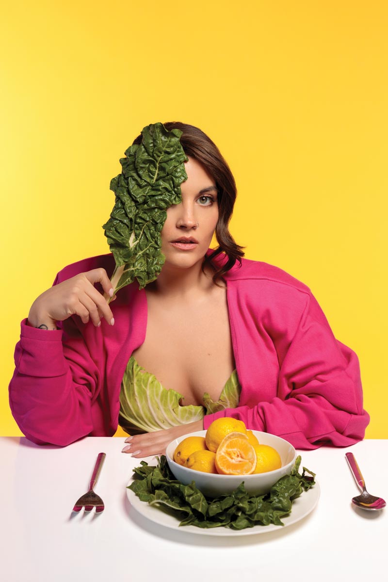 woman sitting at table with plate of food in front of her holding a kale leaf up to her face, on a bright yellow background