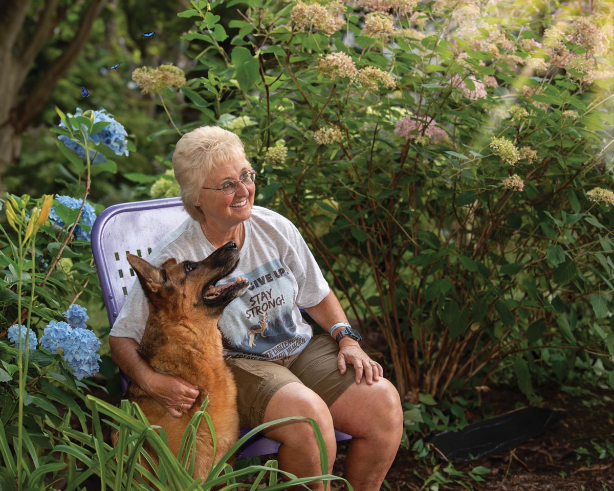 woman sitting in a chair in a lush garden with her arm around her german shepherd dog, who is looking up at her smiling