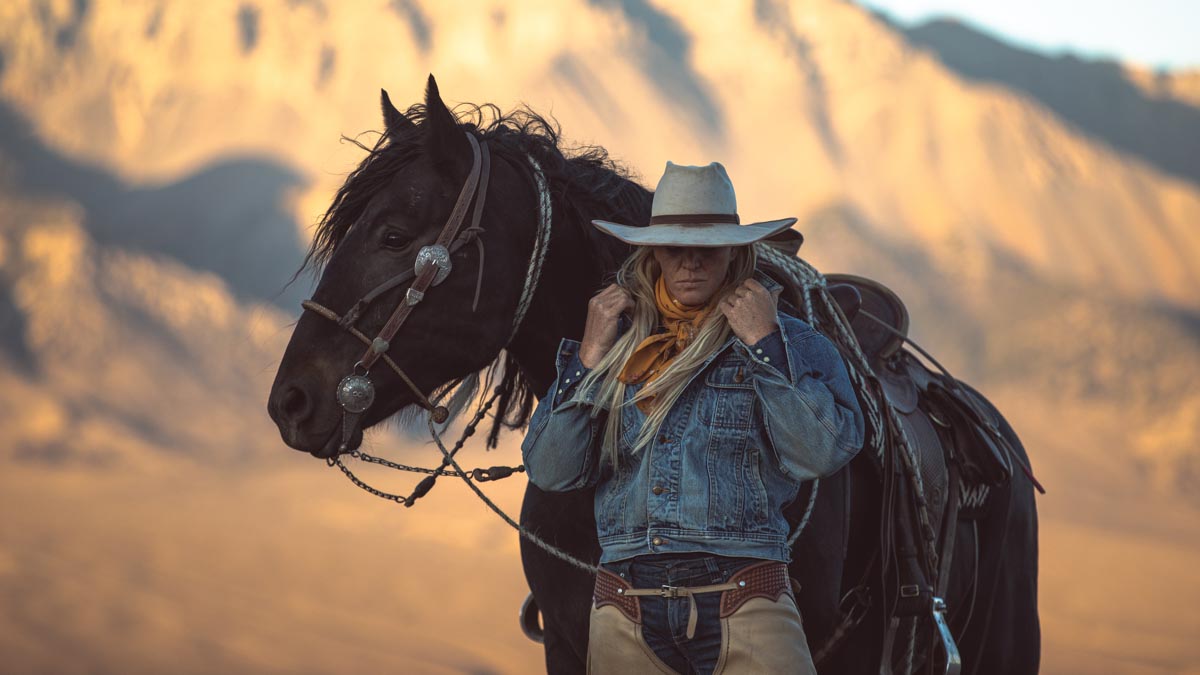 woman wearing cowboy hat standing with horse with mountains in background
