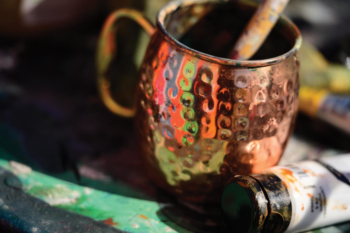 A copper cup typically used for a Moscow Mule sits with a paint brush resting in it and a tube of paint next to it on a table