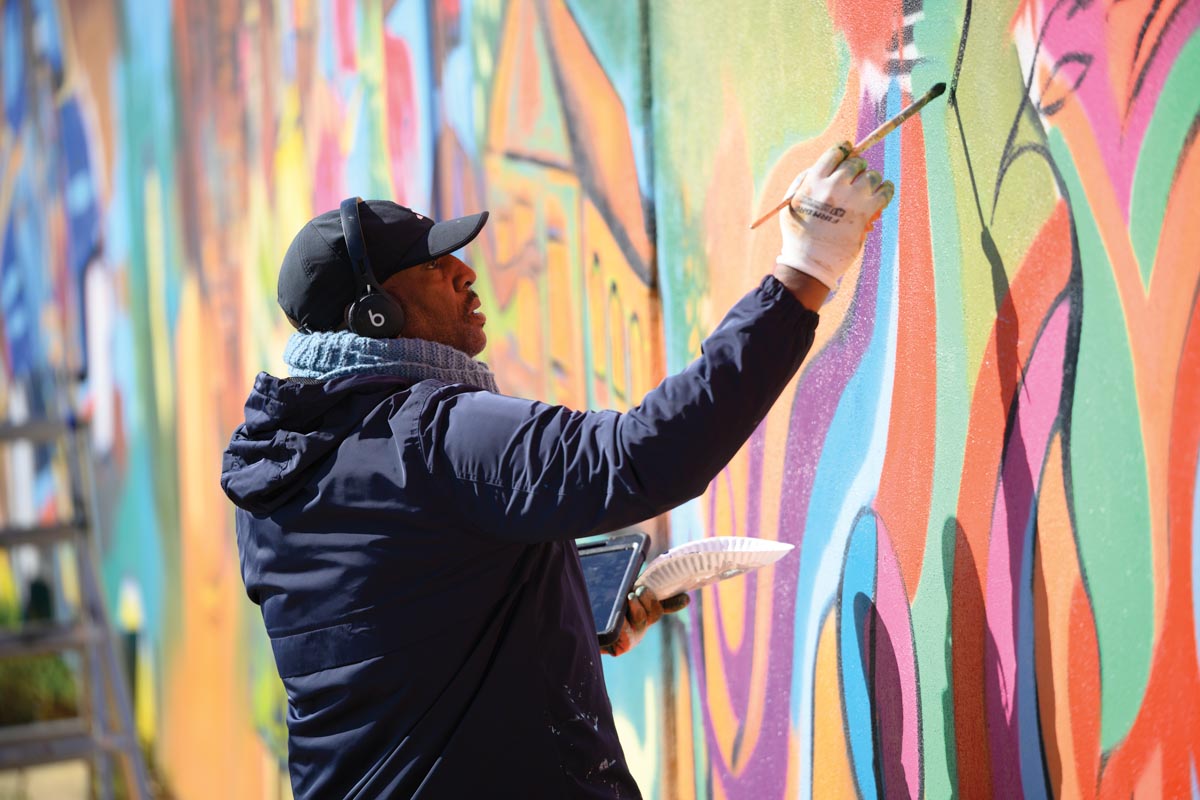 A painter wearing a black hat, earphones, and a hooded jacket works on a mural with a long paint brush