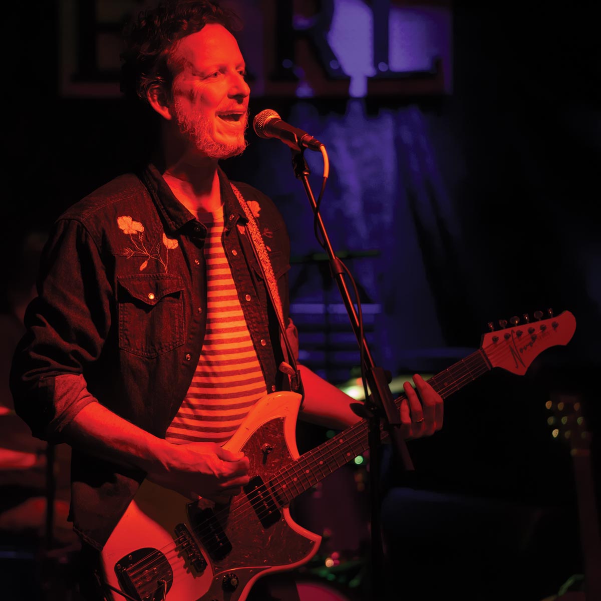 A bearded guitarist performs under red light wearing a denim shirt over a horizontally striped shirt