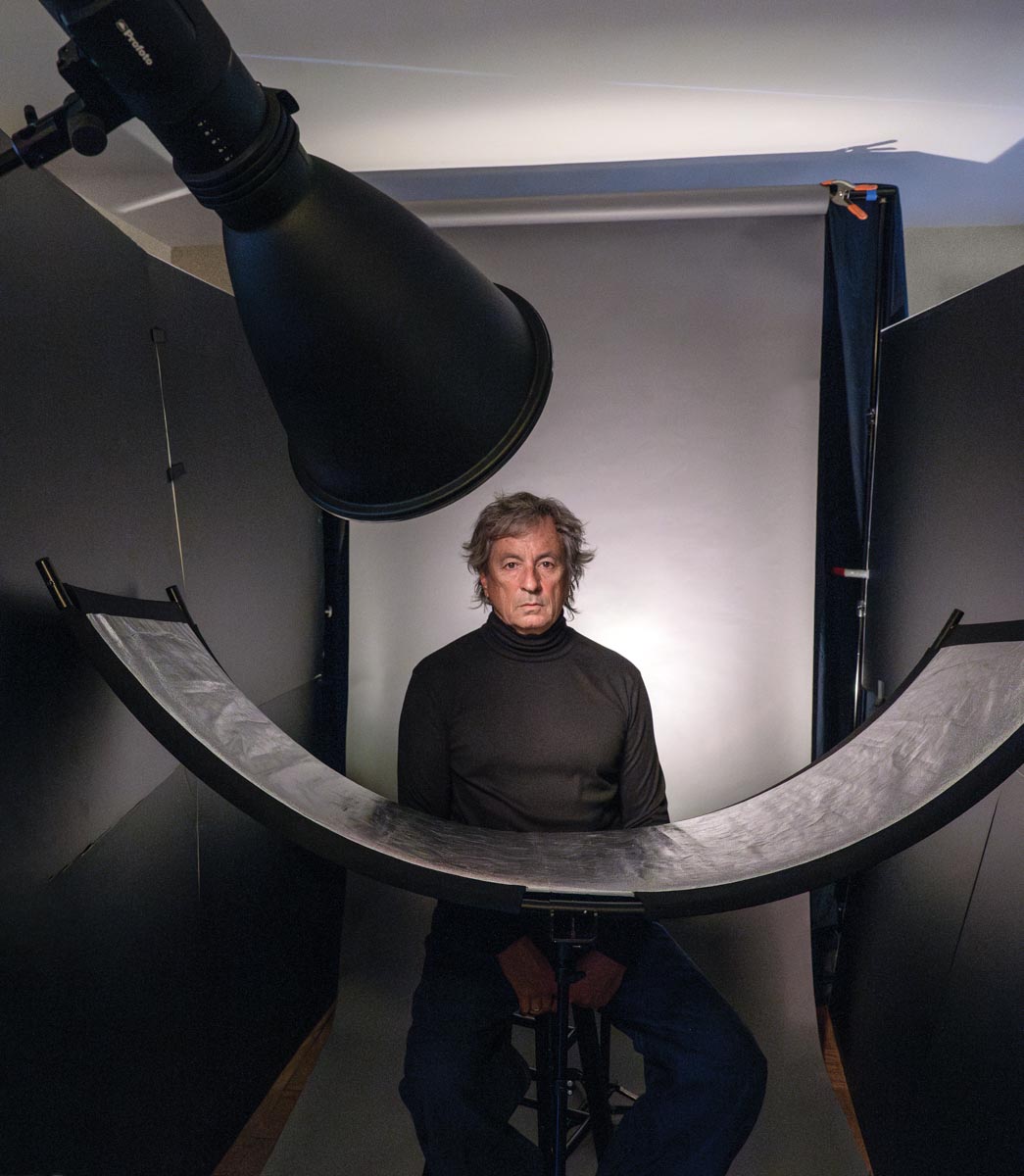 A man sits for a portrait with a backdrop and v-flats behind him and a Westcott Eyelighter below and in front of his chest and a light modified with a hard reflector in the foreground