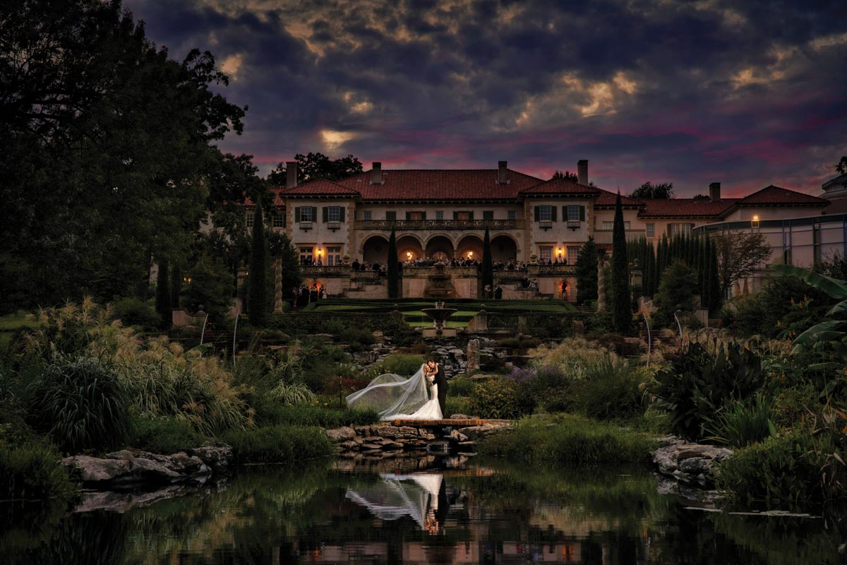 bride and groom in front of a pond with a grand hotel with a Spanish tile roof and cloudy sky in the background