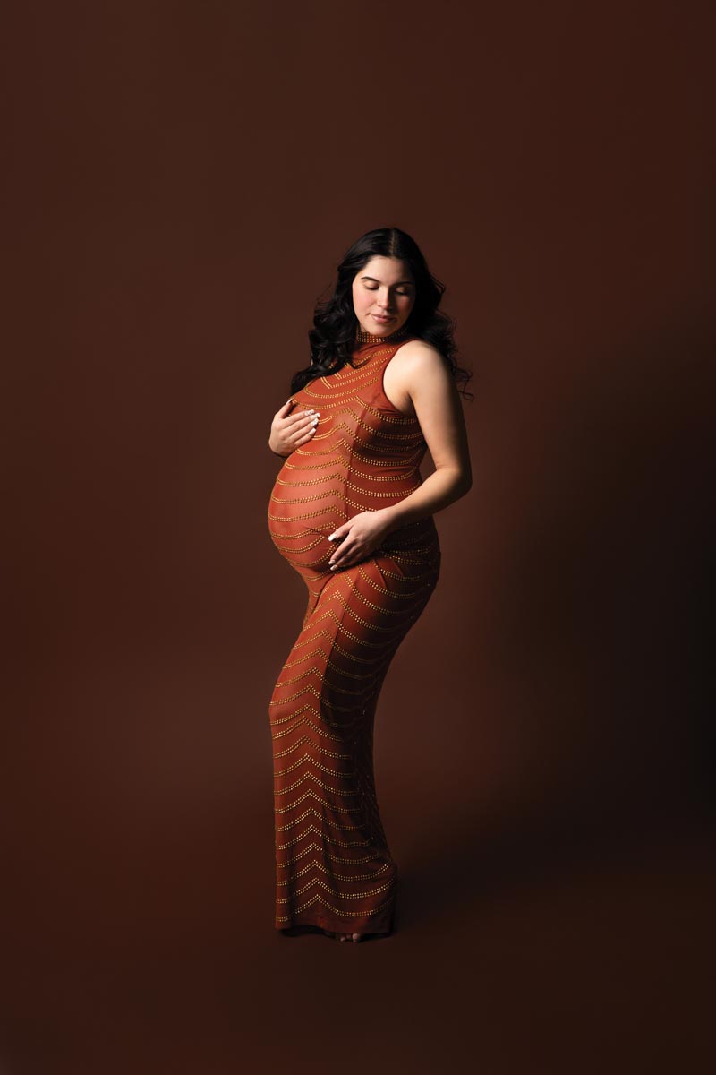 pregnant woman in front of dark orange backdrop wearing a burnt orange down showing off her baby bump