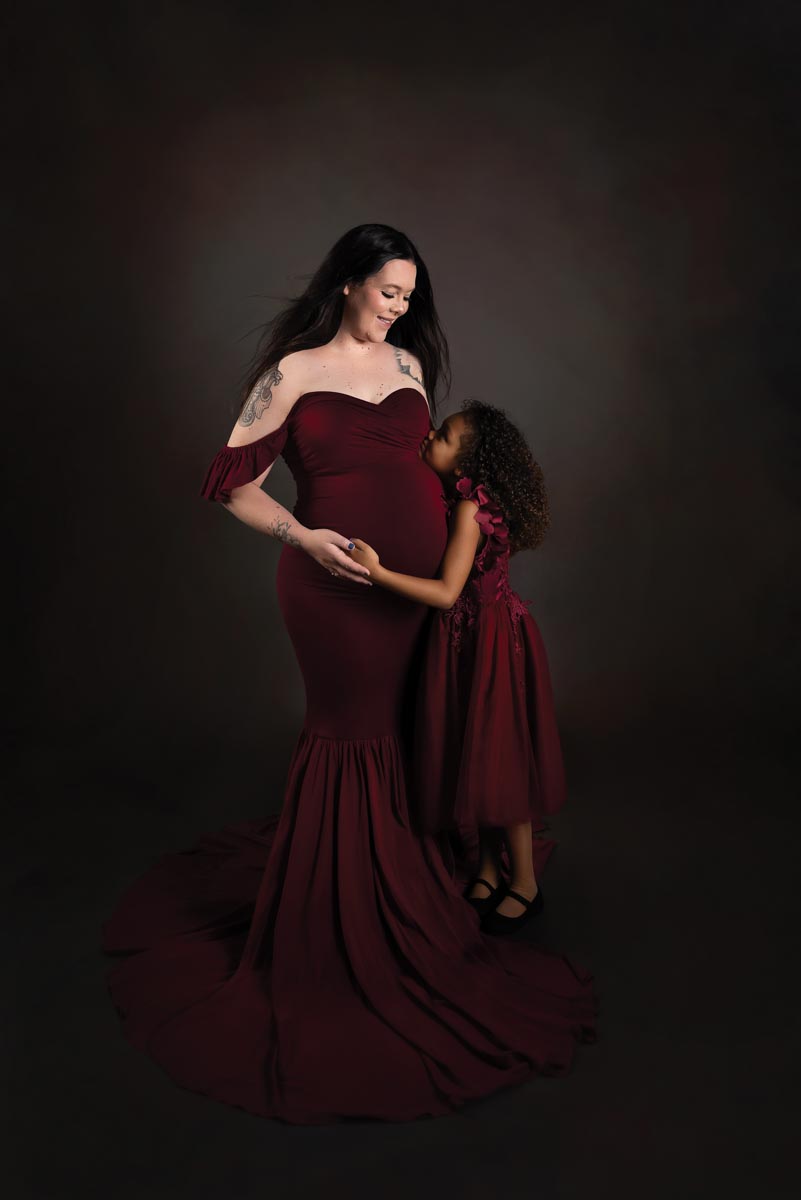 pregnant woman in maroon gown looking down at her young daughter whose arm is touching her baby bump