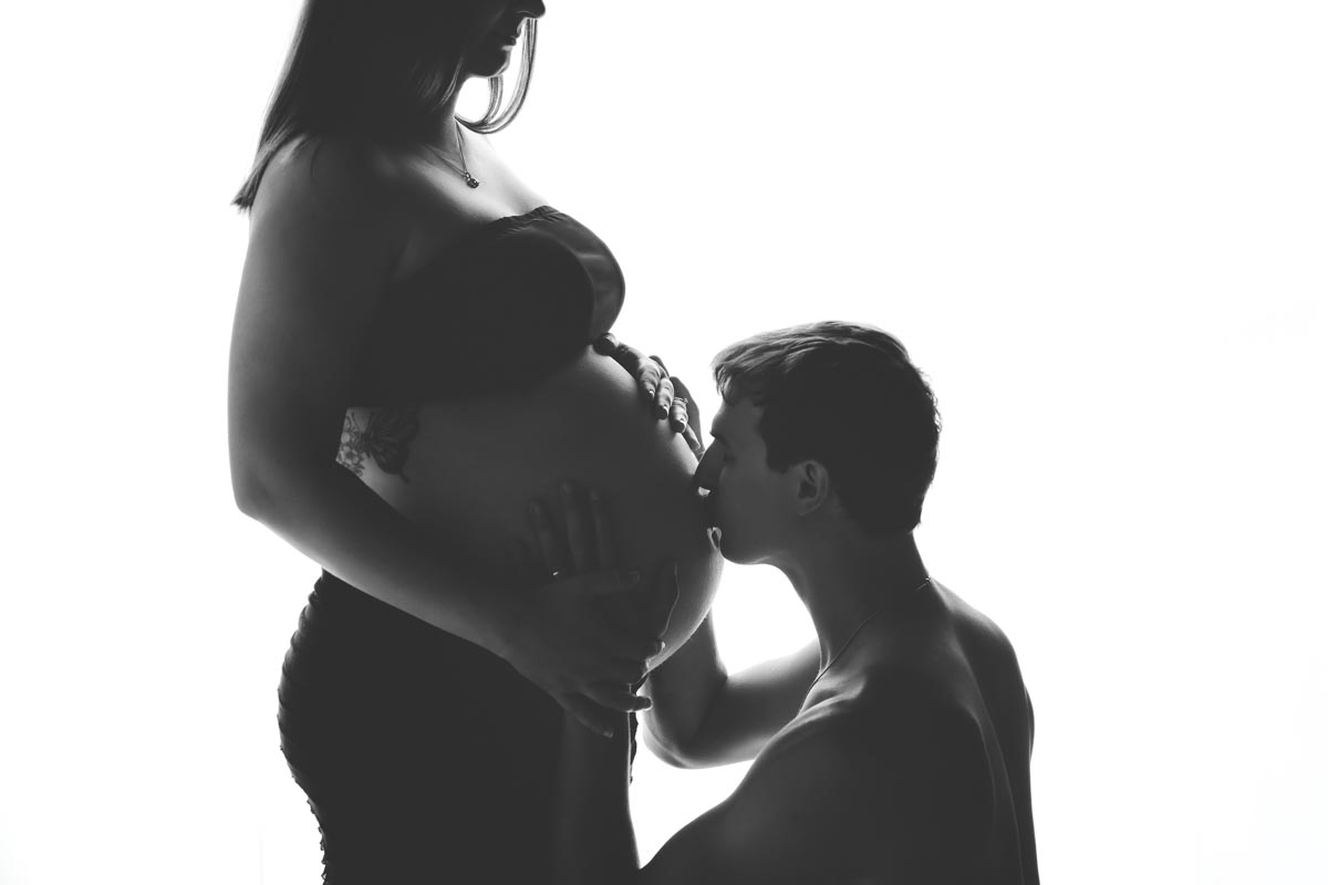 black-and-white photo of pregnant mother with bare baby bump that father is kneeled down to kiss