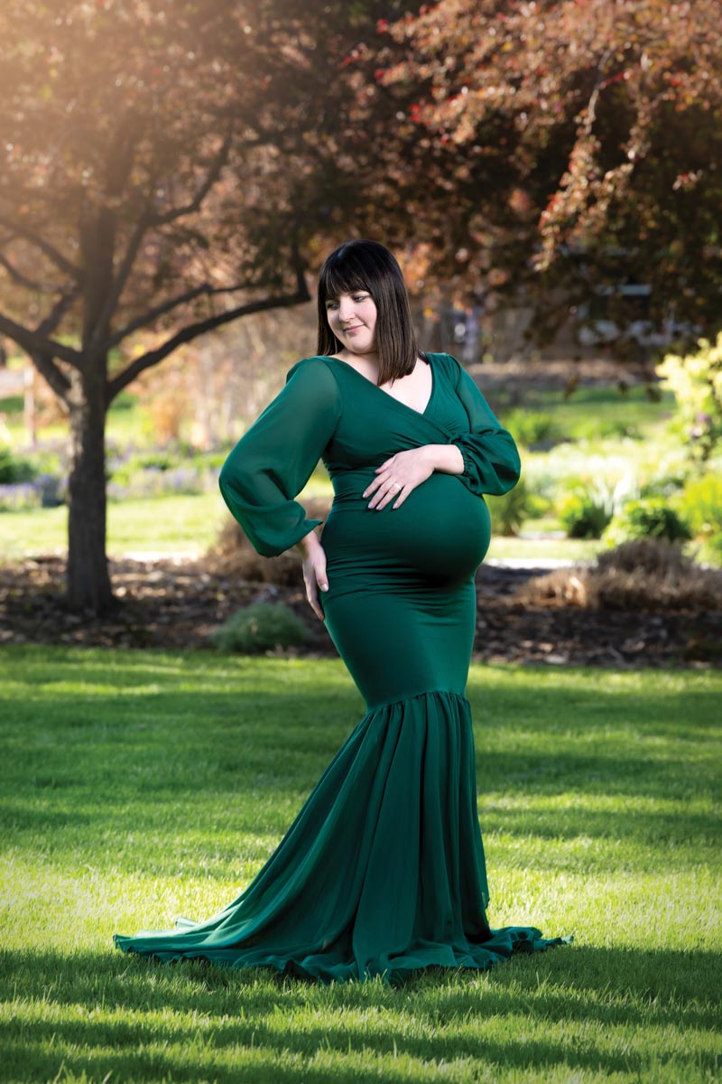 pregnant woman wearing a green gown standing in a grassy field with trees in the background