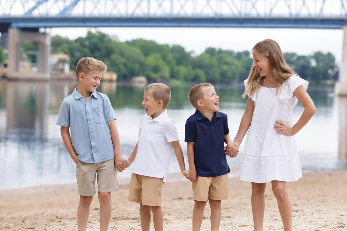 photo of four young children holding hands and looking at each other on a beach with a bridge in the background 