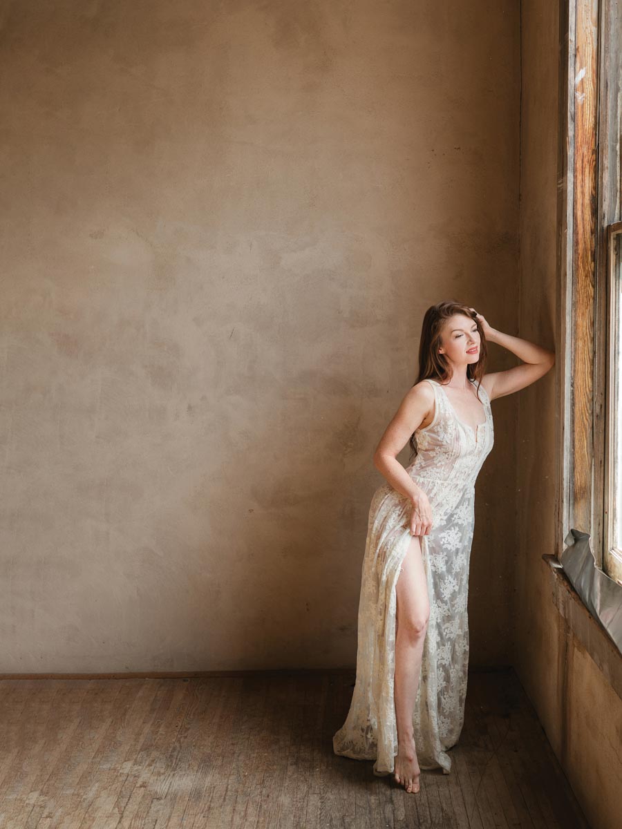 woman standing near window wearing a lace dress in front of a beige wall