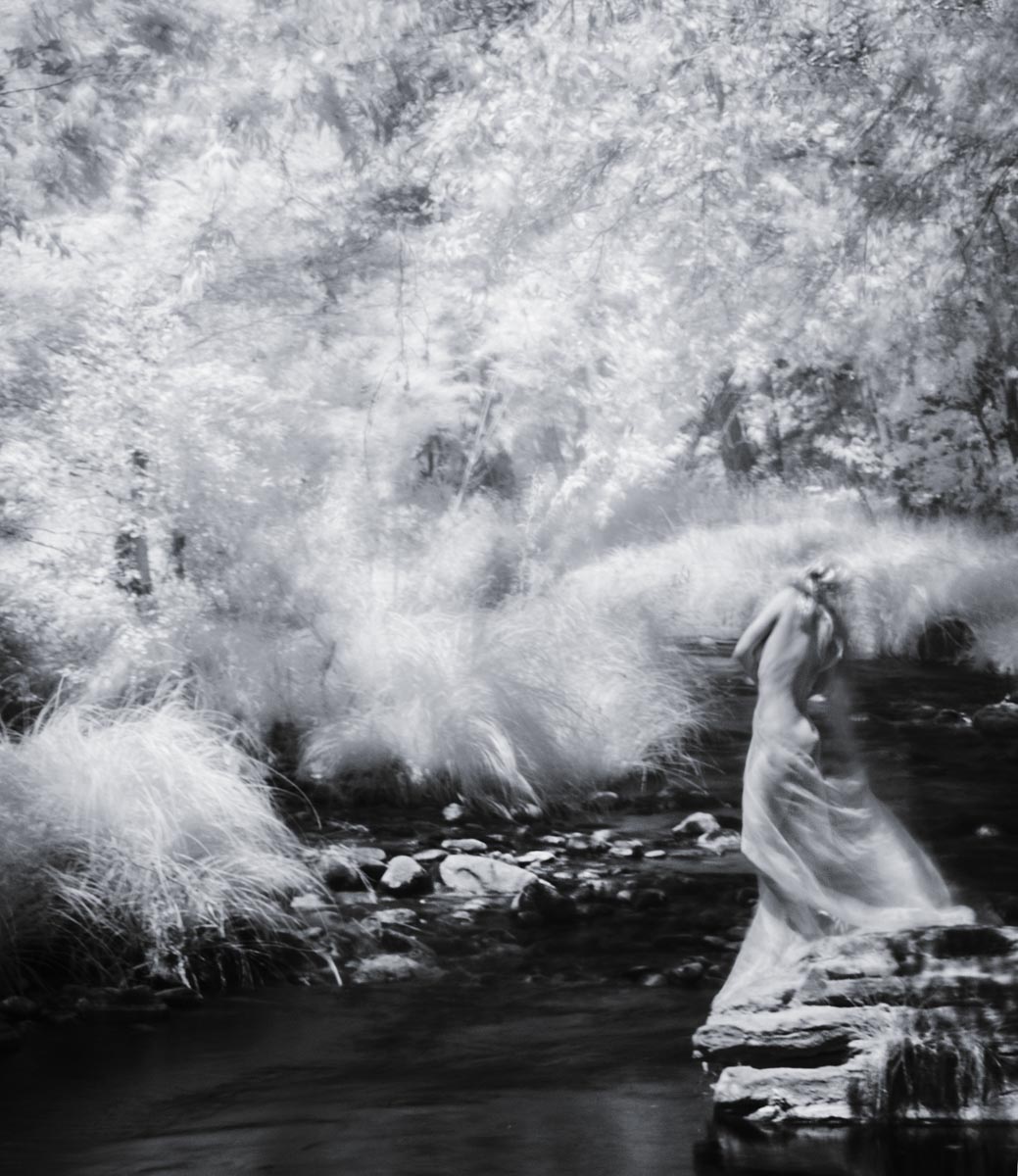 infrared photo of a femal figure standing at the edge of a river near trees