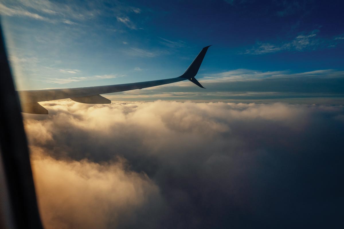 a starboard airplane wing as seen through the airplane window as it flies just above the clouds toward sunset
