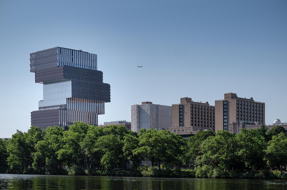 A building that looks like stacks of metal rises into the sky on the left, and three brown apartment buildings on the right, a faint airplane in the sky, trees separate the buildings from a body of water 