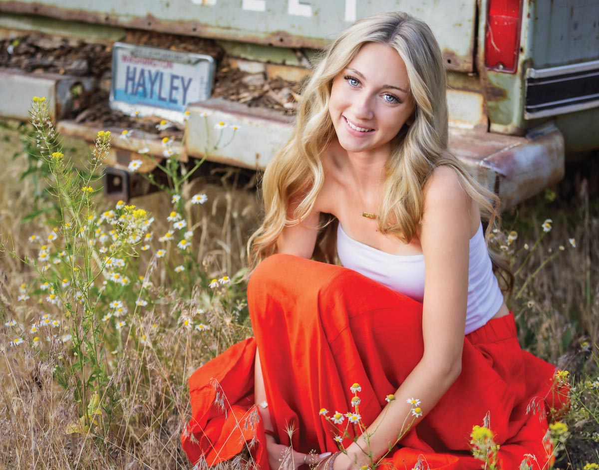 Blonde girl in a red skirt and white tank top sits on grass next to the back of a car and smiles at the camera