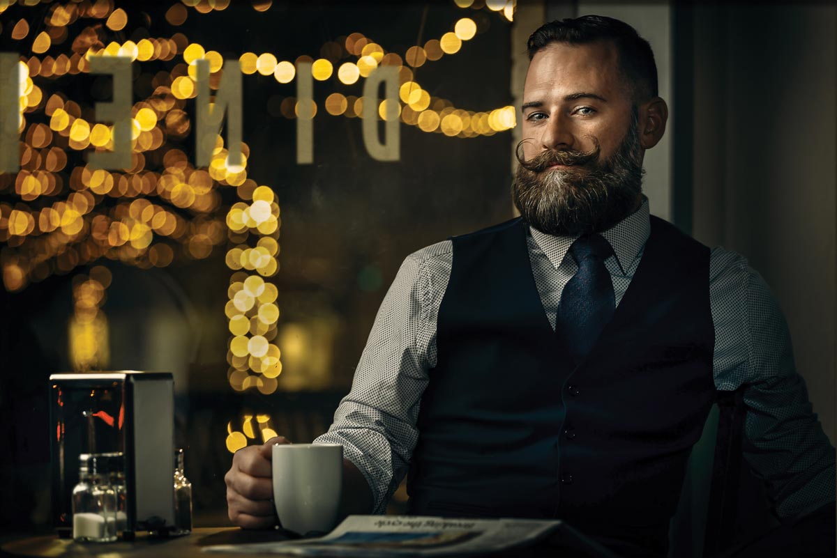 A man with a full beard and mustache in a suit with a vest, holding a mug with the light fixture of a restaurant behind him