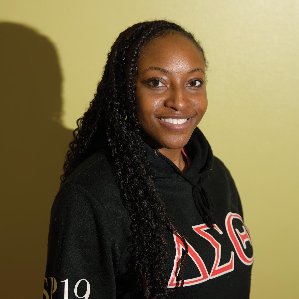 A Black woman with braids wearing a Delta Sigma Theta hoodie, in front of a yellow background. Light from one direction throws a sharp shadow behind her, and there is a little light glare on her face. 