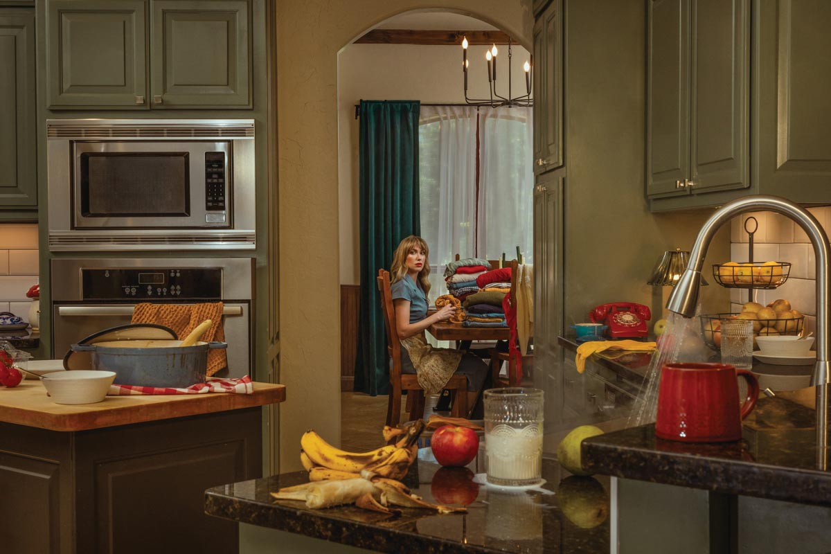 Alex Eischeid conceptual self-portrait of her sitting in the dining room looking into a messy kitchen